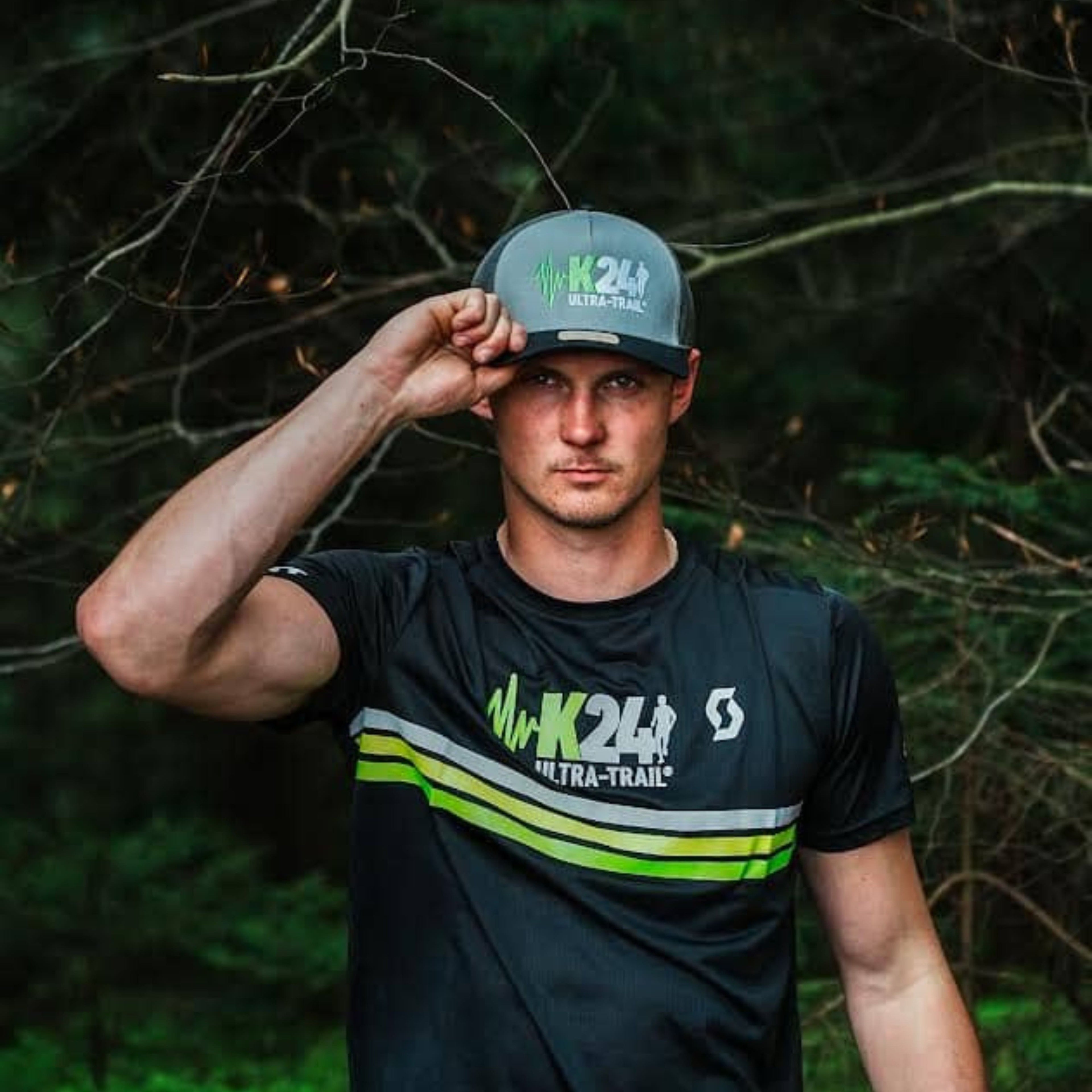 Man wearing a black t-shirt with 'K24 Ultra-Trail' logo and a cap in a forest setting
