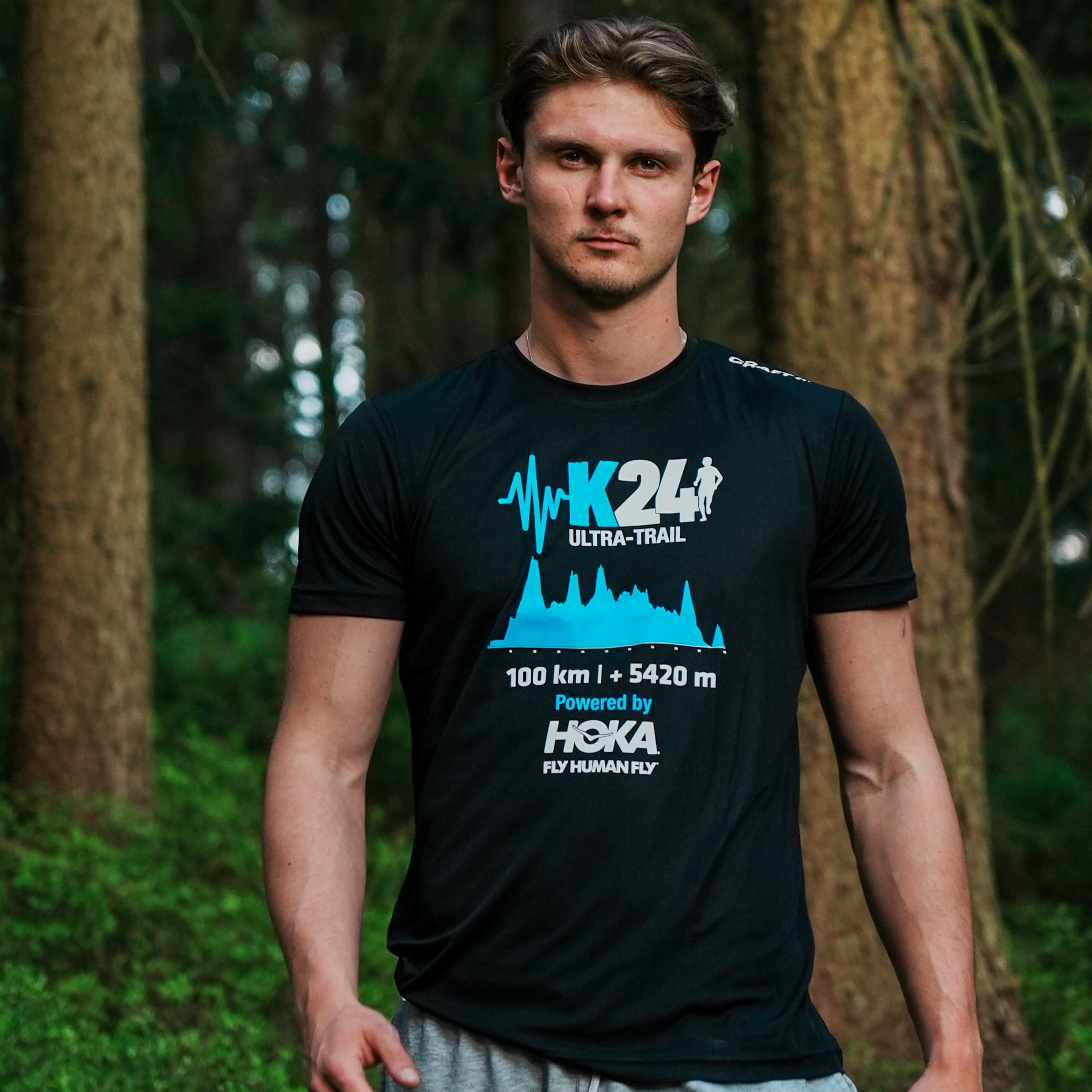 Man wearing a black t-shirt with blue and white graphics and text, standing in a forest.