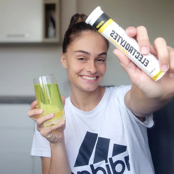 Person holding a glass of yellow liquid and a tube labeled 'Electrolytes' in a kitchen setting.