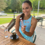 Woman holding a supplement tube outdoors at a table with a drink and water glass.