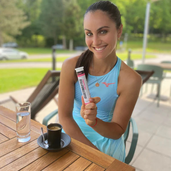 Woman holding a supplement tube outdoors at a table with a drink and water glass.