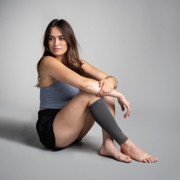 Woman sitting on a gray background wearing a gray sleeveless top and black shorts with a gray compression calf sleeve.