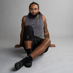 Man sitting on a gray background wearing a gray tank top and black knee brace with an orange logo.