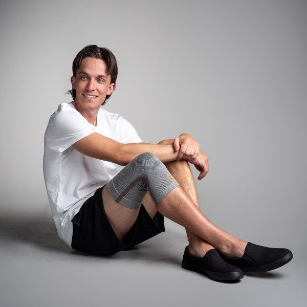Man sitting on a gray background wearing a white shirt, black shorts, and gray knee sleeves.