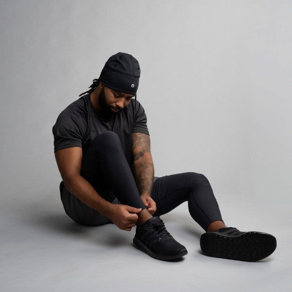 Man sitting on the floor wearing black long pants, tying his black shoes against a gray background.