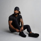 Man sitting on the floor wearing black clothing and shoes against a gray background