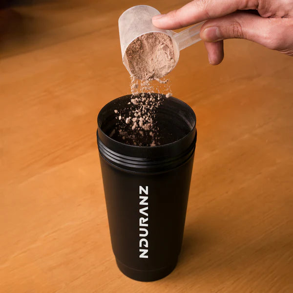 Person adding powder from a scoop into a black NDURAN cup on a wooden surface